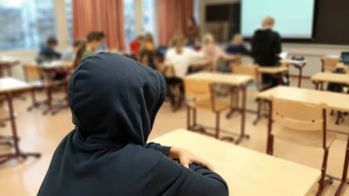 En elev sitter med huvan uppdragen på sin mörkblå munkjacka, långt borta från den övriga klassen.