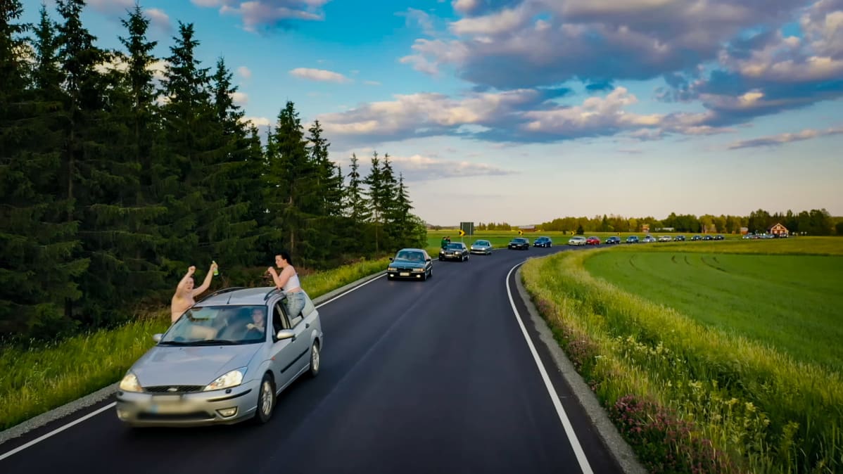 Kaksi henkilöä istuu auton ikkunoiden ulkopuolella auton kulkiessa autoletkan ensimmäisenä.