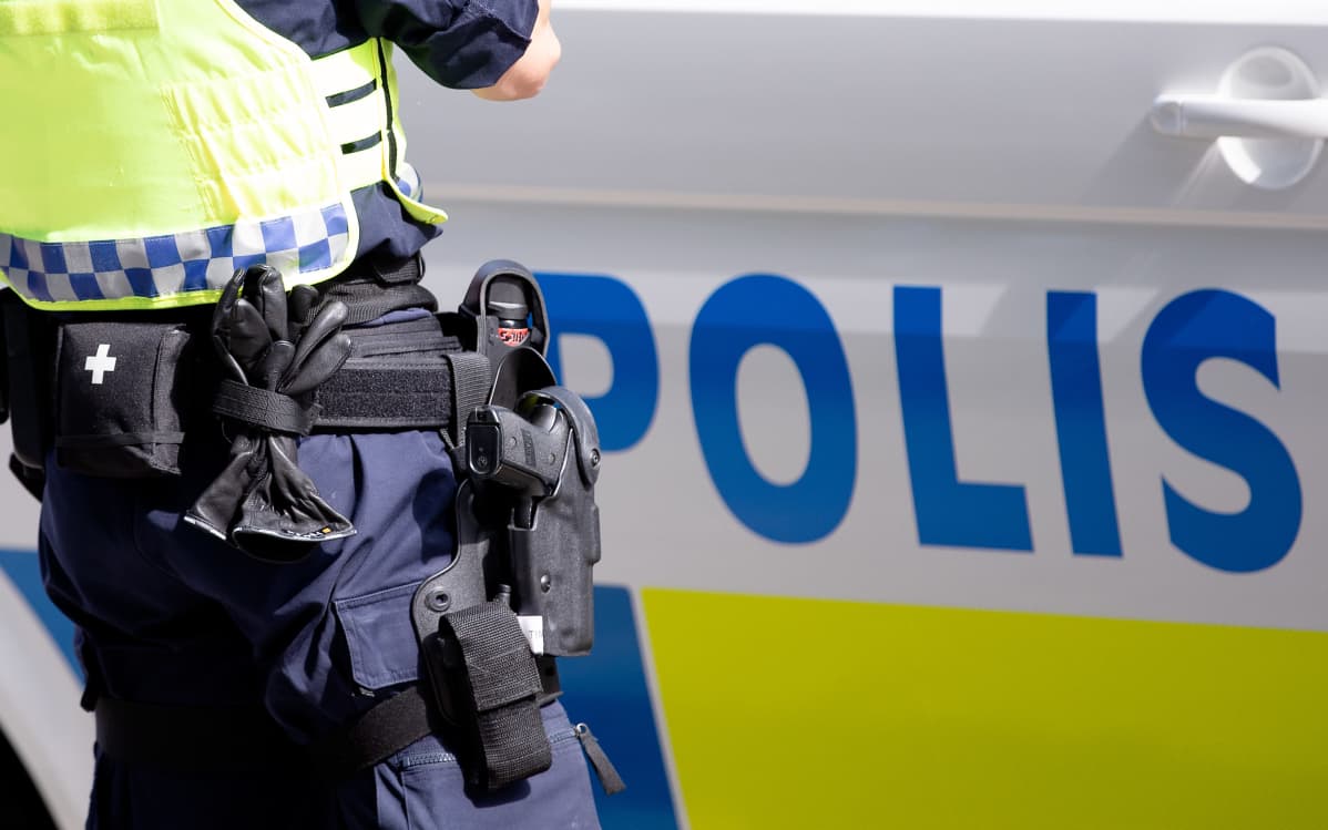 Poliisi seisoo pistooli vyöllään poliisiauton vieressä.