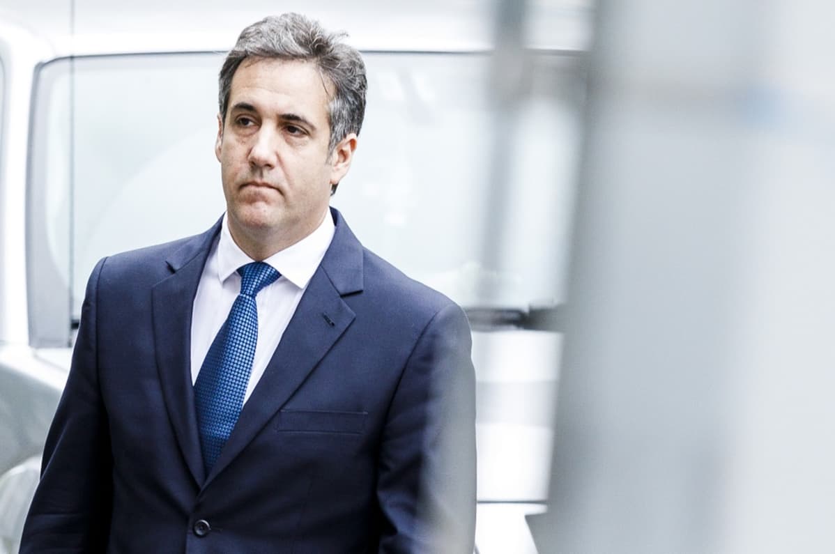 Cohen kävelee tummansinisessä puvussa, sininen kravatti kaulassaan, vakava ilme kasvoillaan.