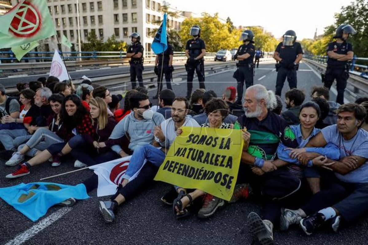 Kuvassa protestoijia istumassa kadulla. Heidän takanaan seisoo poliiseja.