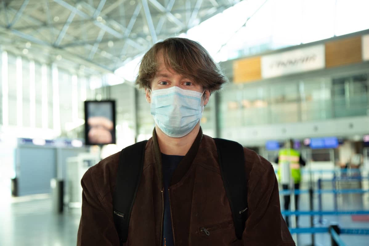 Dylan Pashly Helsinki-Vantaa lentoaseman lähtöaulassa. Dylanilla on ruskea takki ja kasvoilla hengityssuojain.