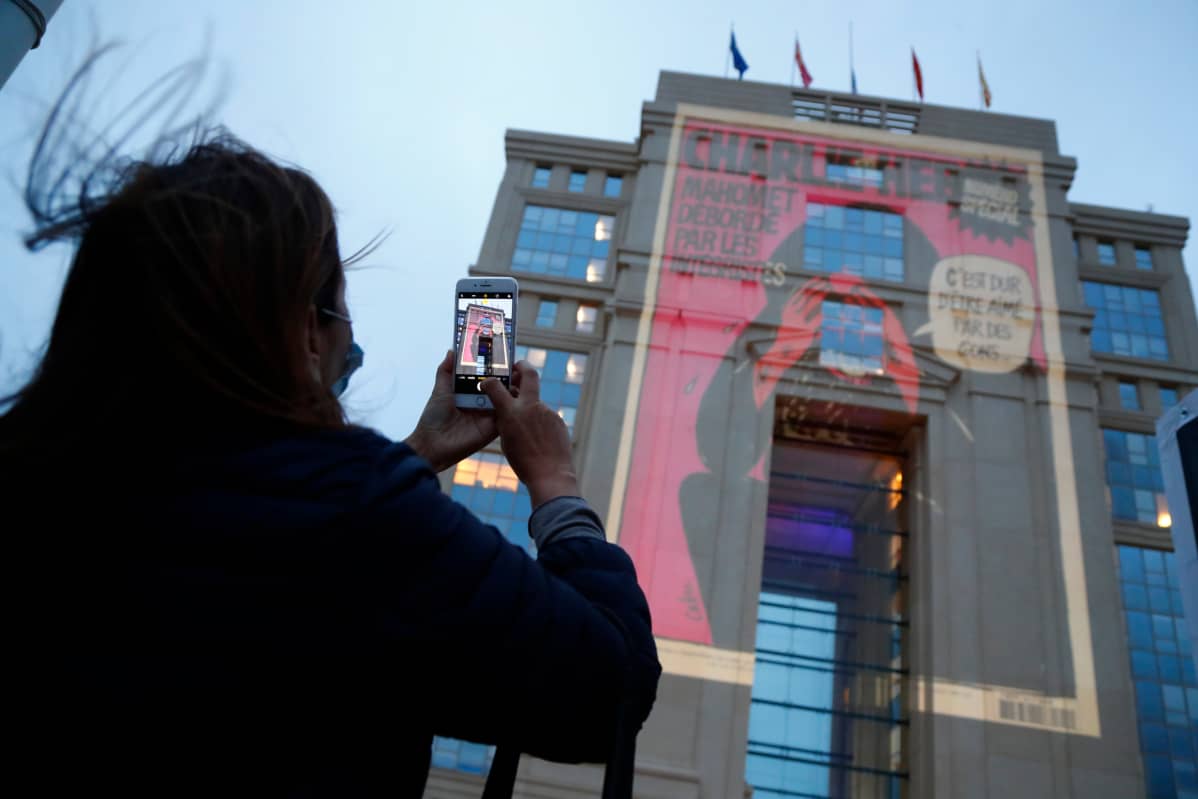 A woman takes pictures with her mobile phone as cartoons from French satirical newspaper Charlie Hebdo are projected onto buildings in central Montpellier in France, 21 October 2020. The cartoons of Prophet Mohammed were projected to pay tribute to the teacher Samuel Paty who was assassinated in Conflans-Sainte-Honorine. On 16 October French school teacher Samuel Paty was decapitated by 18-year-old attacker named Abdoulakh Anzorov who has been shot dead by policemen. Paty was a history teacher who had recently shown caricatures from Charlie Hebdo newspapers of the Prophet Mohammed in class.