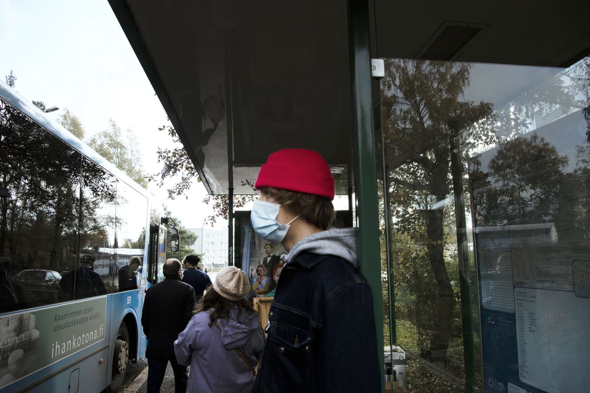 Kasvomaskiin pukeutunut nuori bussipysäkillä.