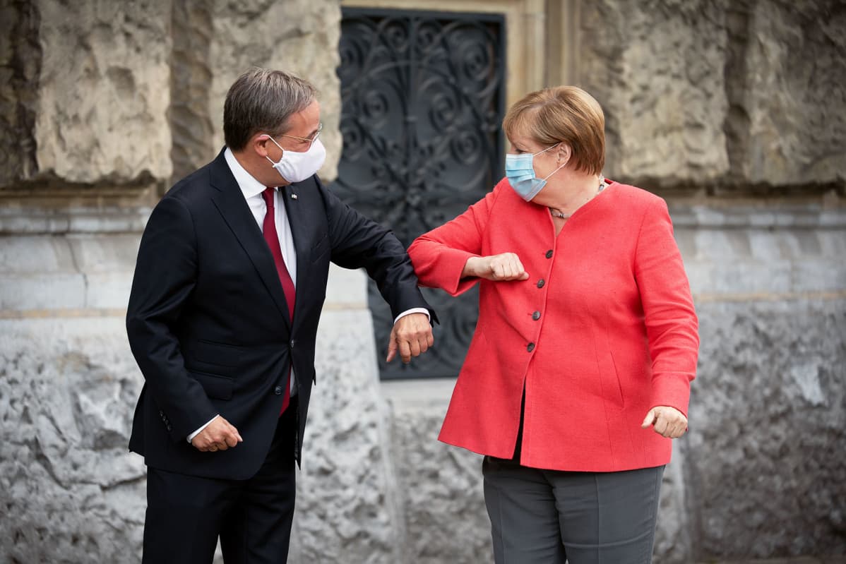 KAsvomaskeihin pukeutuneet Armin Laschet ja Angela Merkel kyynärtervehtivät toisiaan.