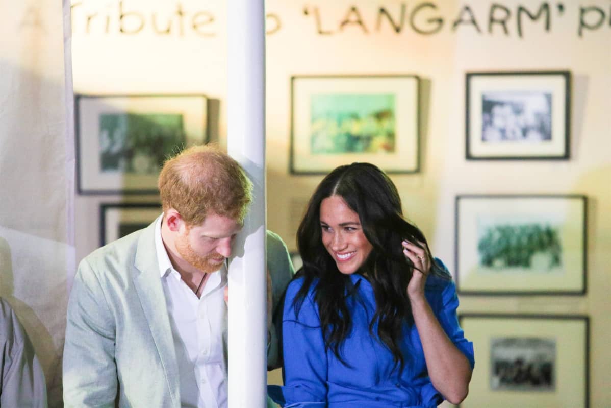 Pylvääseen nojaava prinssi Harry ja hymyilevä Meghan.