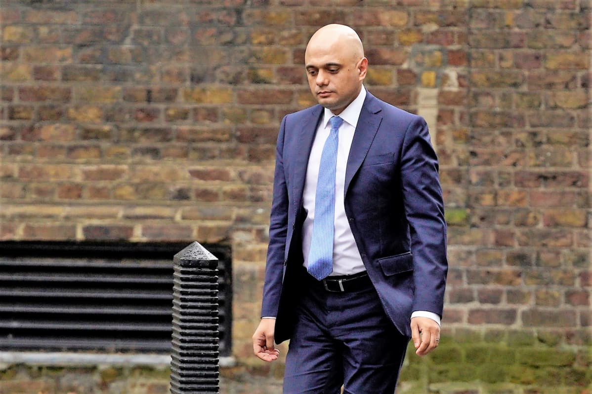 Sajid Javid kävelee tiiliseinän edustalla. Hänellä on sininen puku ja vaaleansininen kravatti.