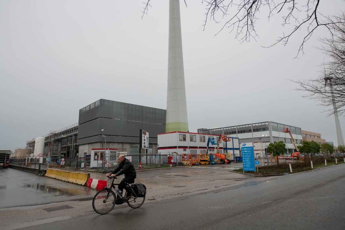 Tuotantolaitoksen rakennuksia, korkea piippu, etualalla pyöräilijä