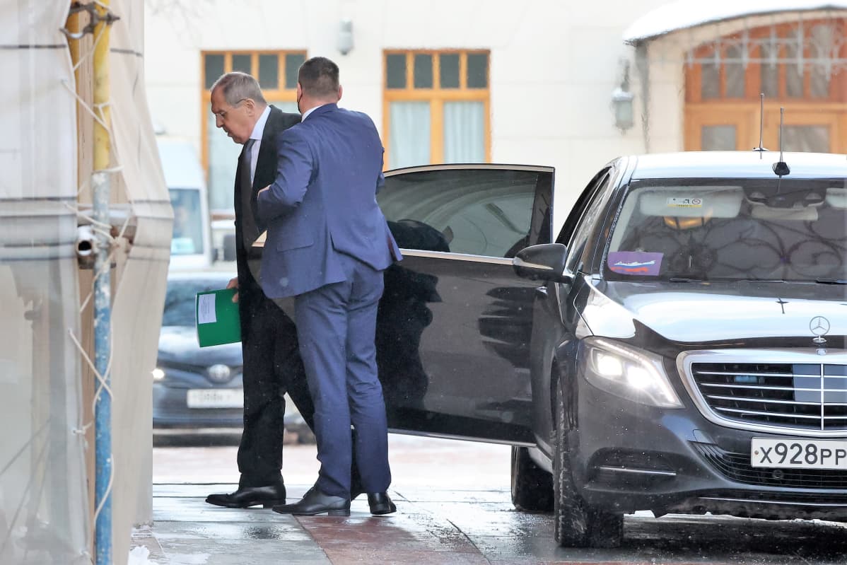 Lavrov on tulossa jalkakäytävän viereen pysähtyneestä mustasta autosta. Hänellä on tumma puku ja kravatti. Auton ovea pitelee auki mies sinisessä puvussa.