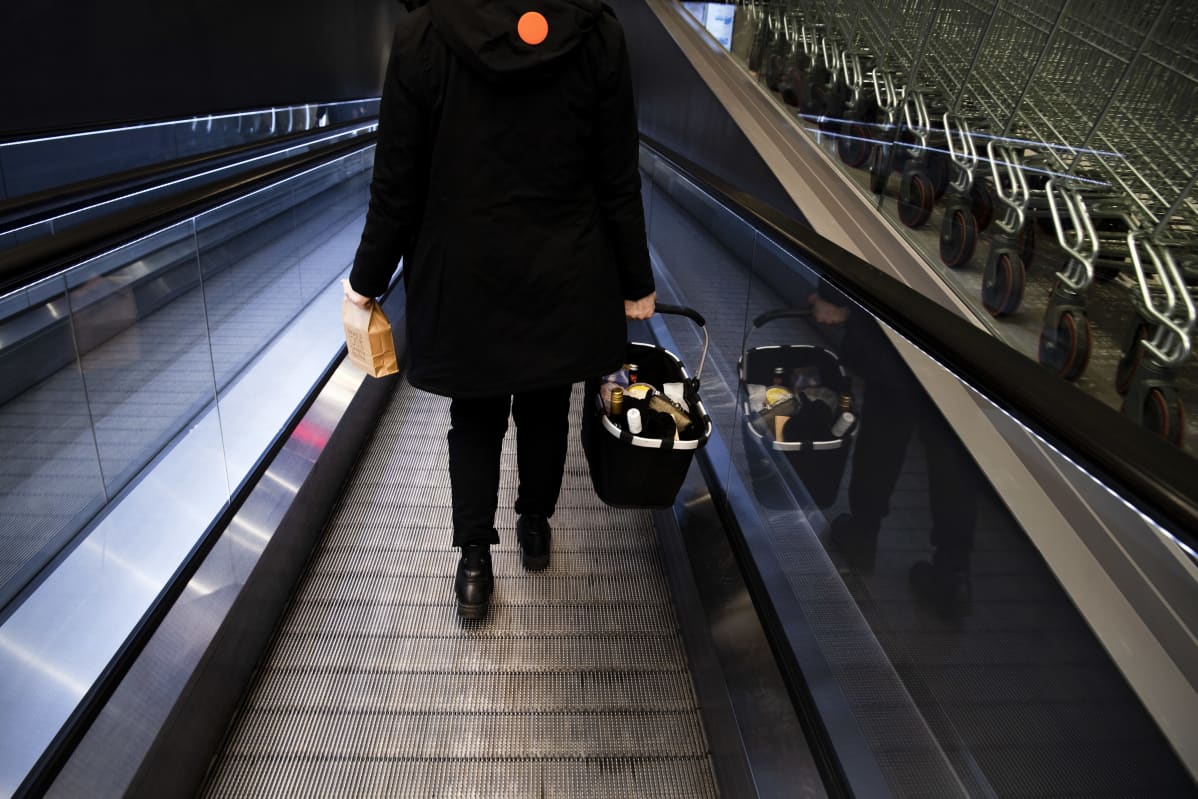 Asiakas kantaa noutoruokapussia ja ostoskoria kauppakeskuksen rullaportaissa.