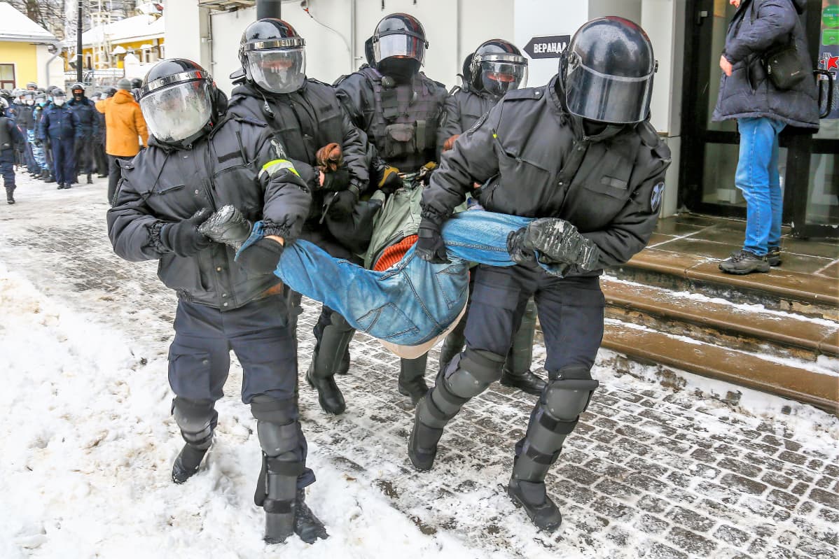 Viisi mellakkapoliisia kantaa keskellään miestä lumisella kadulla. Poliisella on tummat asut ja kypärät visiiri alas laskettuna.