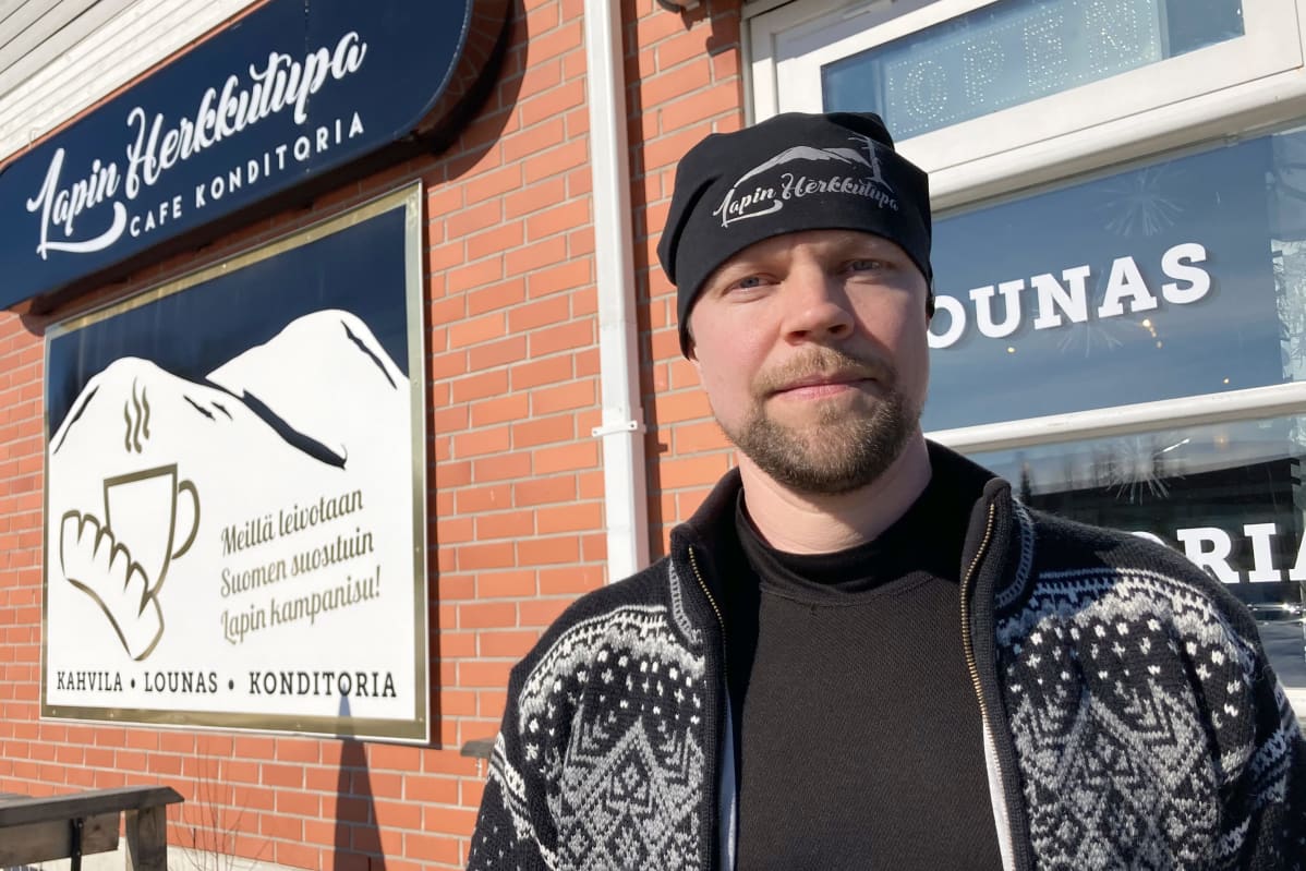 Konditoria-kahvilayrittäjä Matti Hiltunen tummassa neuletakissa. Taustalla tiiliseinää, jossa Hiltusen yrityksen Lapin herkkutuvan mainoskyltti.