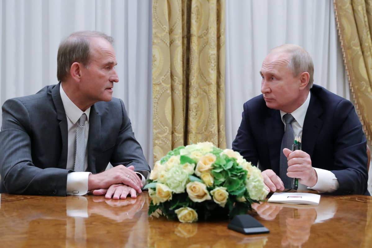 Medvedtshuk ja Putin istuvat pöydän ääressä. Medvedtshuk puhuu ja Putin kuuntelee. Medvedtshuikilla on tummanharmaa puku, Putnilla tumma. Päydällä heidän keskellään on kukka-asetelma.