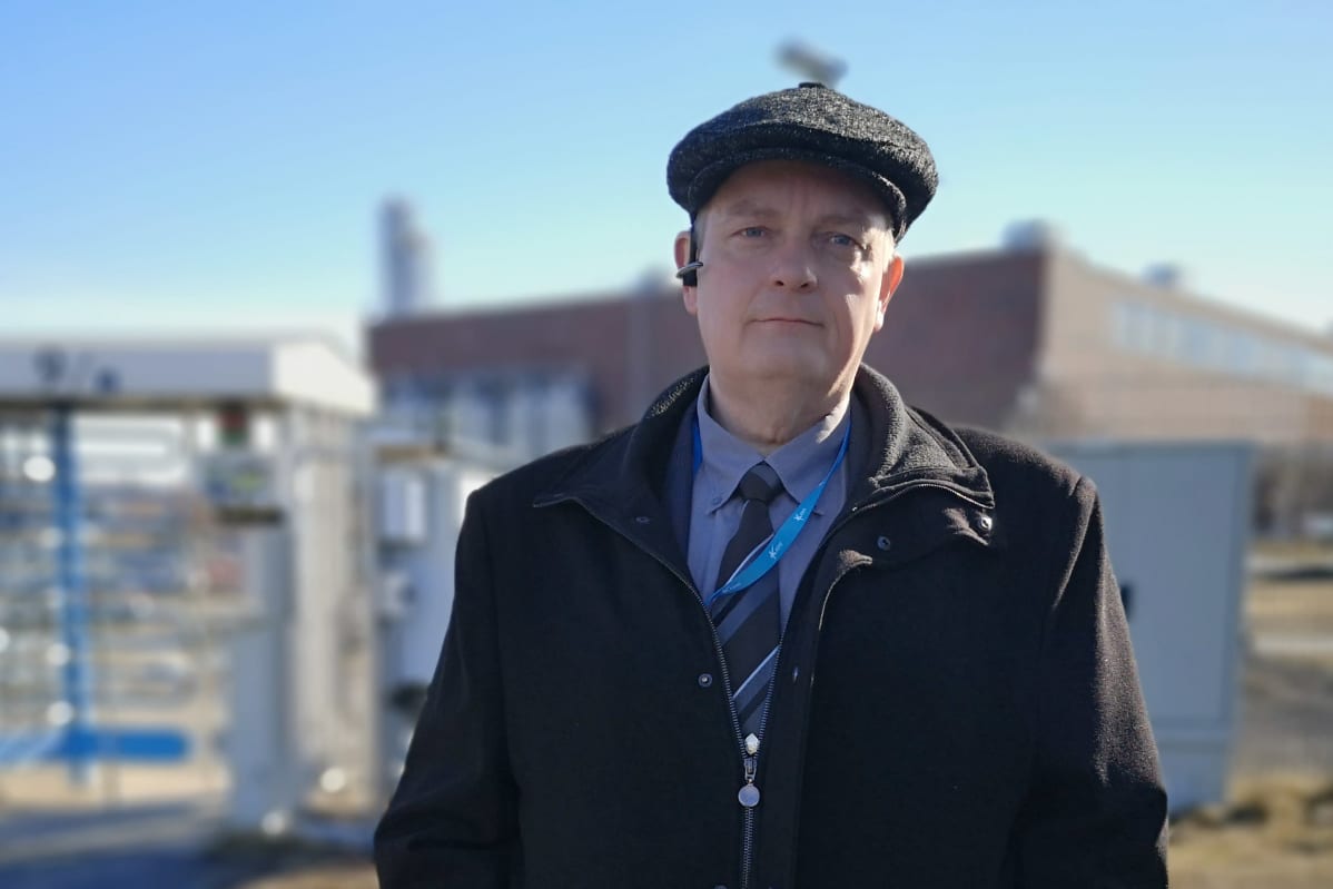 Tummiin pukeutunut Kemin kaupunginjohtaja Matti Ruotsalainen seisoo Veitsiluodon tehtaiden aidan luona, taustalla tehtaan rakennuksia.