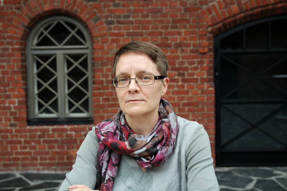 Amnesty Internationalin Suomen osaston ihmisoikeuspuolustajien asiantuntija Anu Tuukkanen punatiilisen seinän edessä, vakava ilme kasvoillaan.
