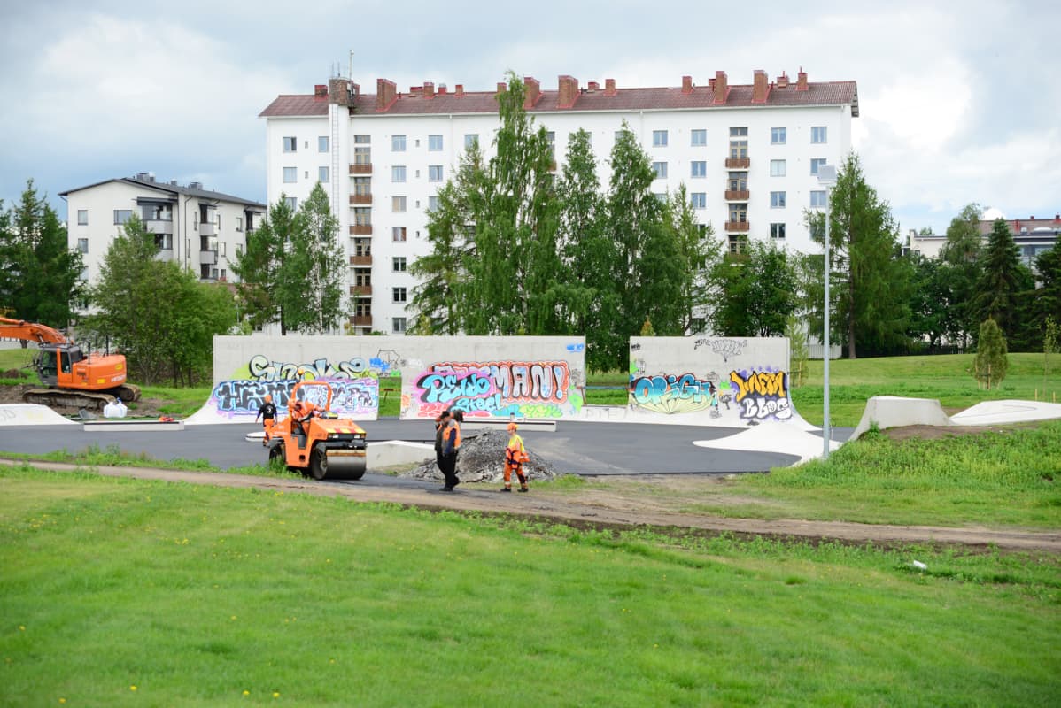 Työmiehiä ja työkoneita Oulun Hollihaan leikkipuistossa, jossa graffitimaalauksia ja taustalla kerrostalo.