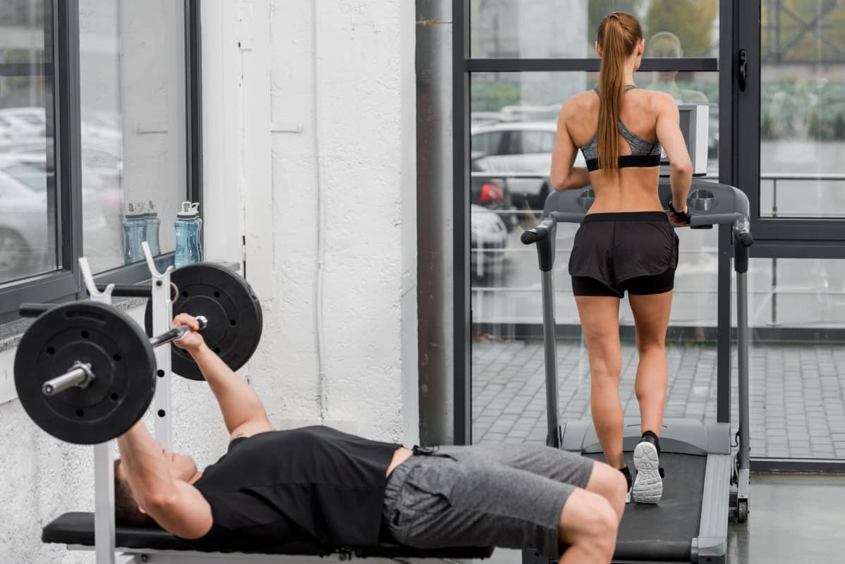 I förgrunden en man som lyfter vikter på gym, i bakgrunden en kvinna som springer på löpmatta.