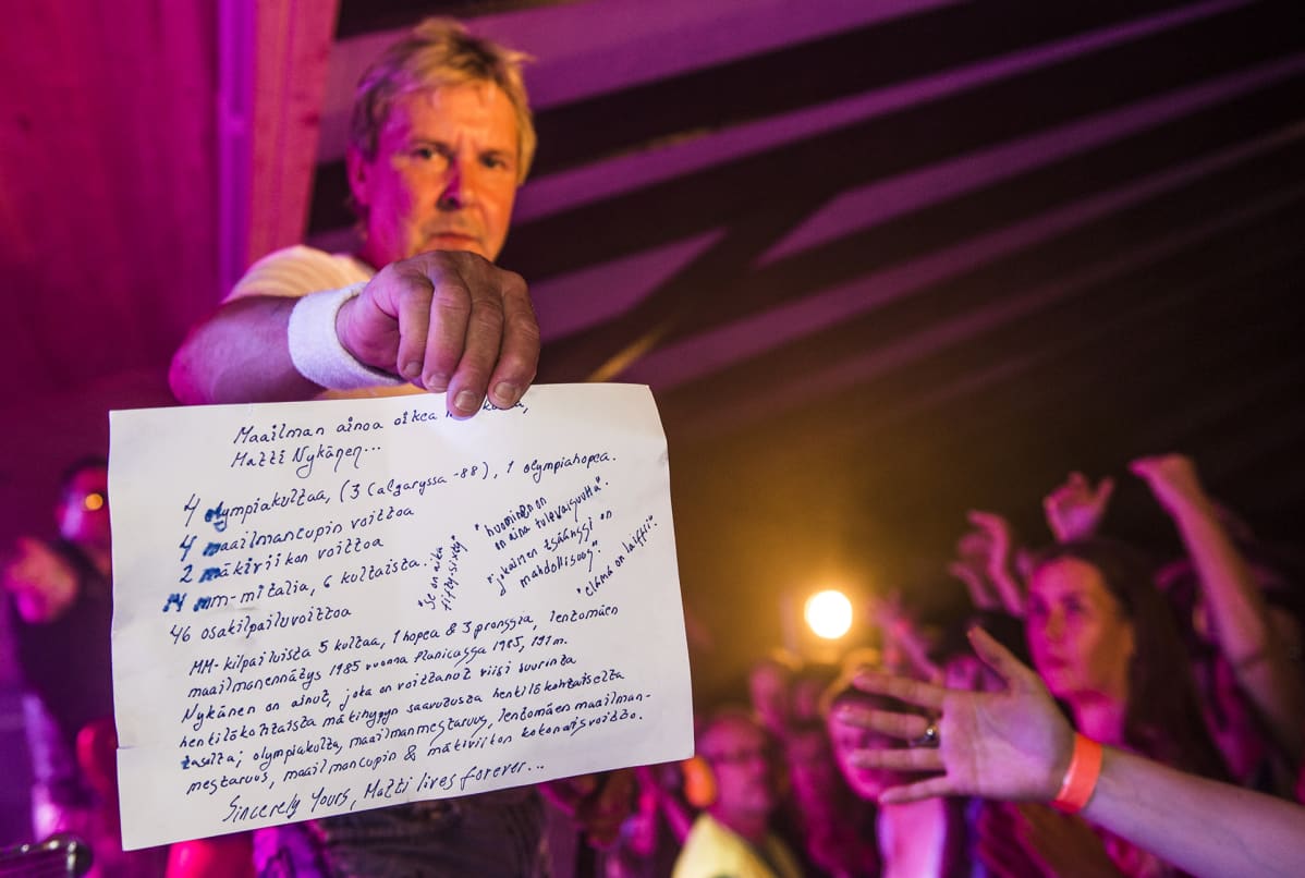 Matti Nykänen näyttää paperia kameralle jossa on lueteltu hänen mitalinsa ja kuolemattomimmat sanonnat.