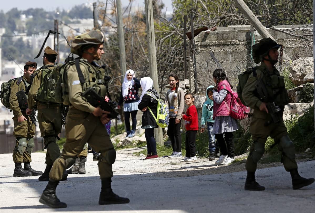 Joukko koulutyttöjä kadun varressa, etualalla neljä raskaasti asiestettua Israelin sotilasta.
