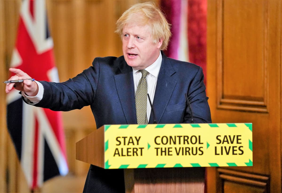Boris Johnson pitää tiedotustilaisuutta tummanisinisessä puvussa, harmaassa kravatissa . Johnson viittoo oikealla kädellään. Hän seisoo puhujanpöntön takana. Pöntössä tekstit: Stay alert. Control the virus. Save lives.