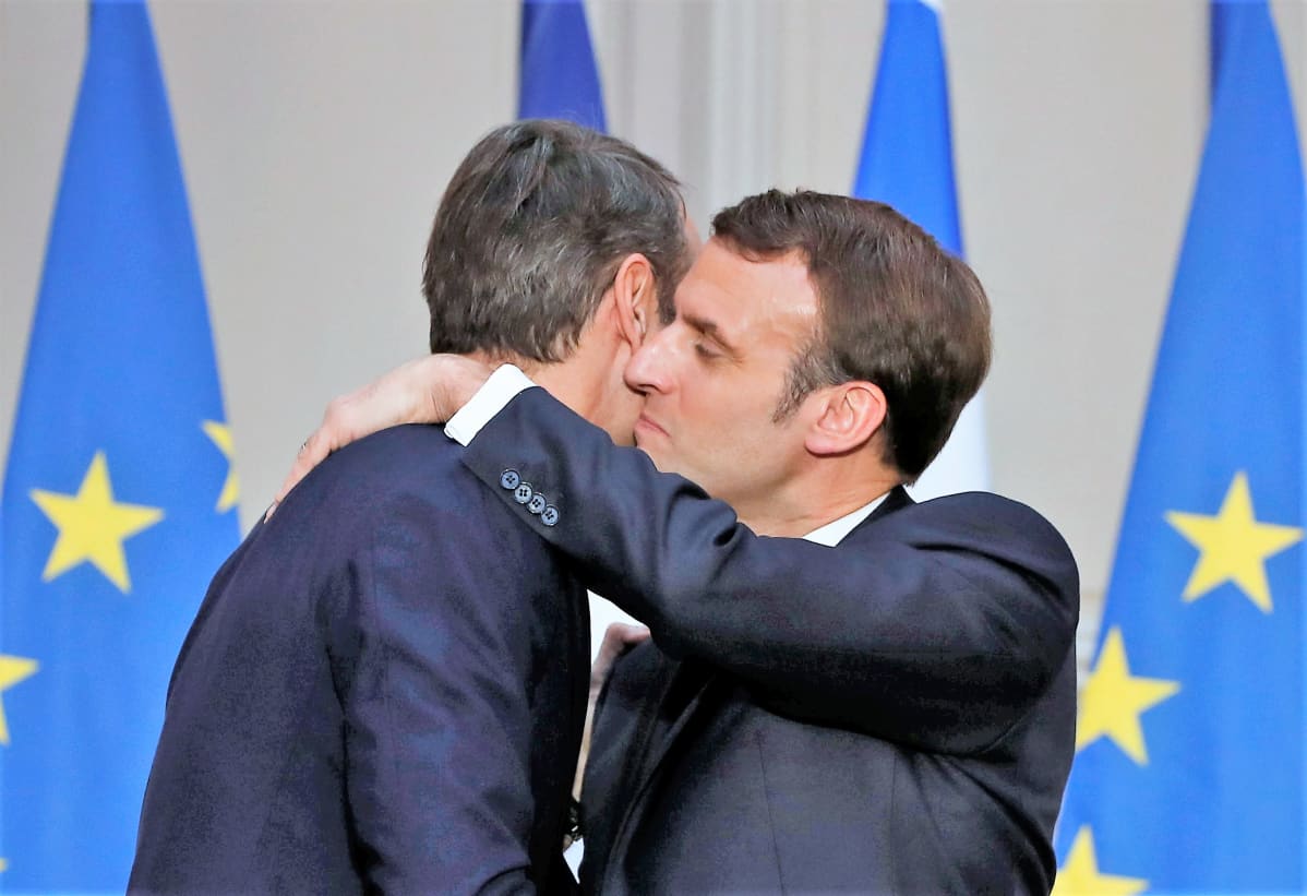 Mitsotakis ja Macron halaavat. Takana näkyy EU-lippuja.