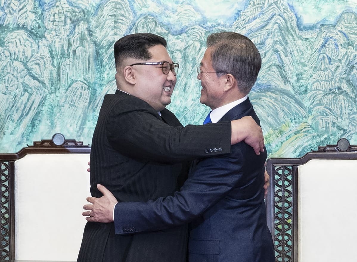 Kim ja Moon halaavat hymyillen kokoushuoneessa. Kimillä on musta maolaishenkinen puku ja Moonilla tummansininen puku.