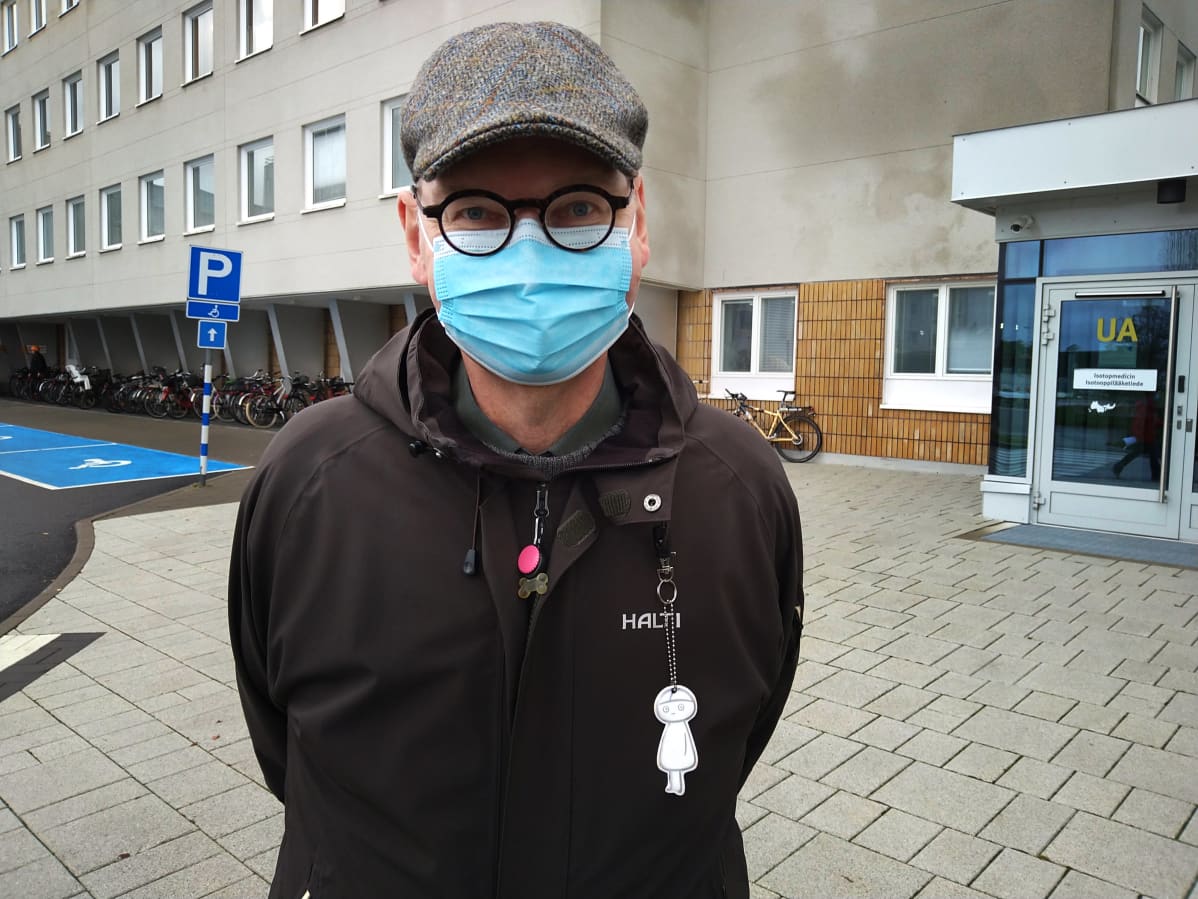Vaasan johtava ylilääkäri Heikki Kaukoranta harmaassa lippalakissa ja tummassa takissa harmaan sairaalarakennuksen edustalla. Kasvoilla on kertakäyttömaski.