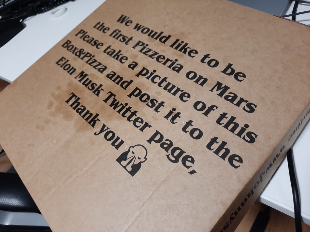 Johnny's Heist pizzalaatikon pohja, jossa lukee englanniksi: Haluamme olla ensimmäinen pizeria Marsissa. Ota kuva tästä tekstistä ja pitsasta. Twiittaa kuvat Elon Muskille.
