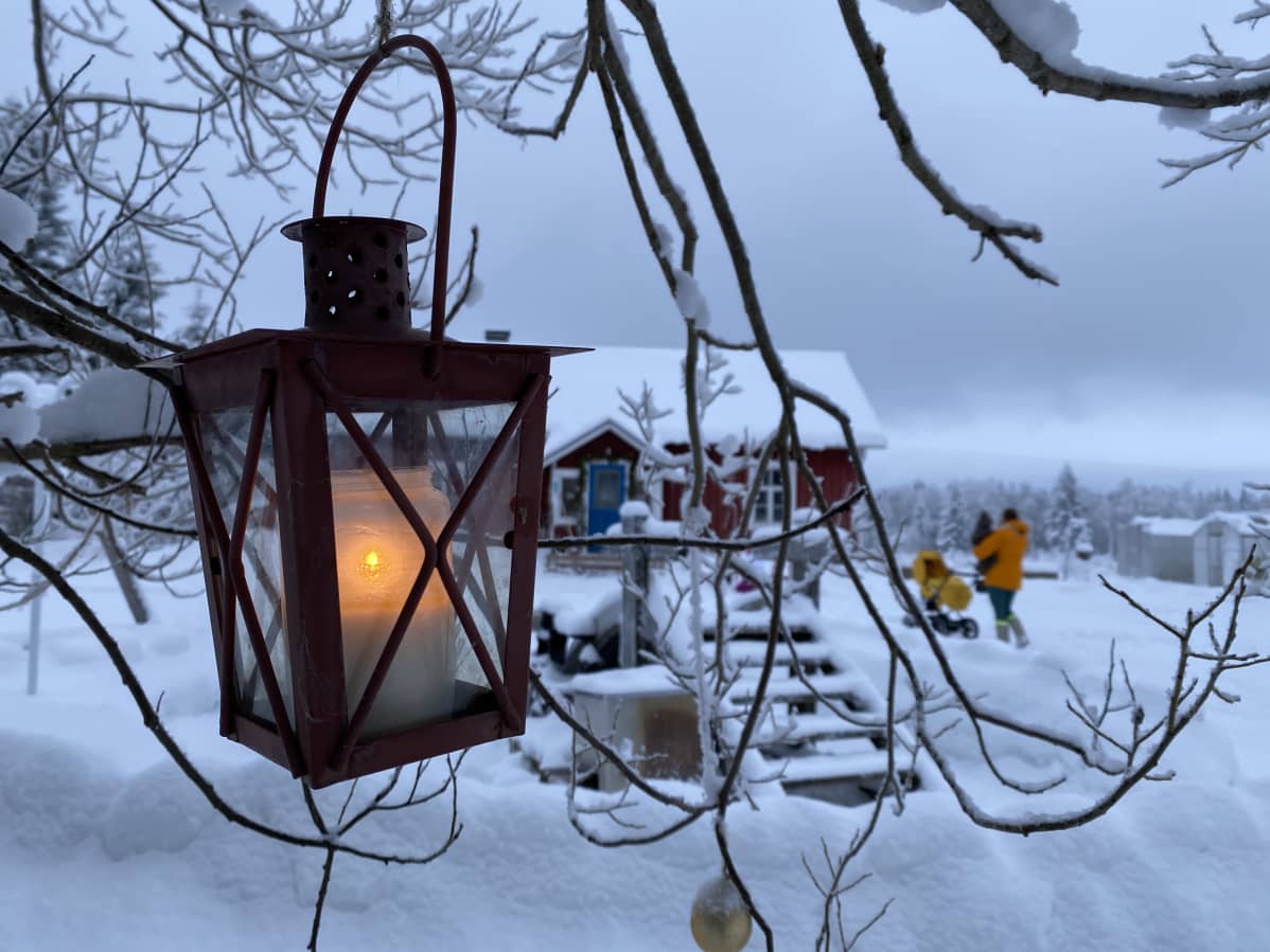 Etualalla lyhty, jossa palaa kynttilä. Se roikkuu puun oksalla. Taustalla punainen hirsitalo. Anne ja lapset ovat kävelemässä kohti taloa.