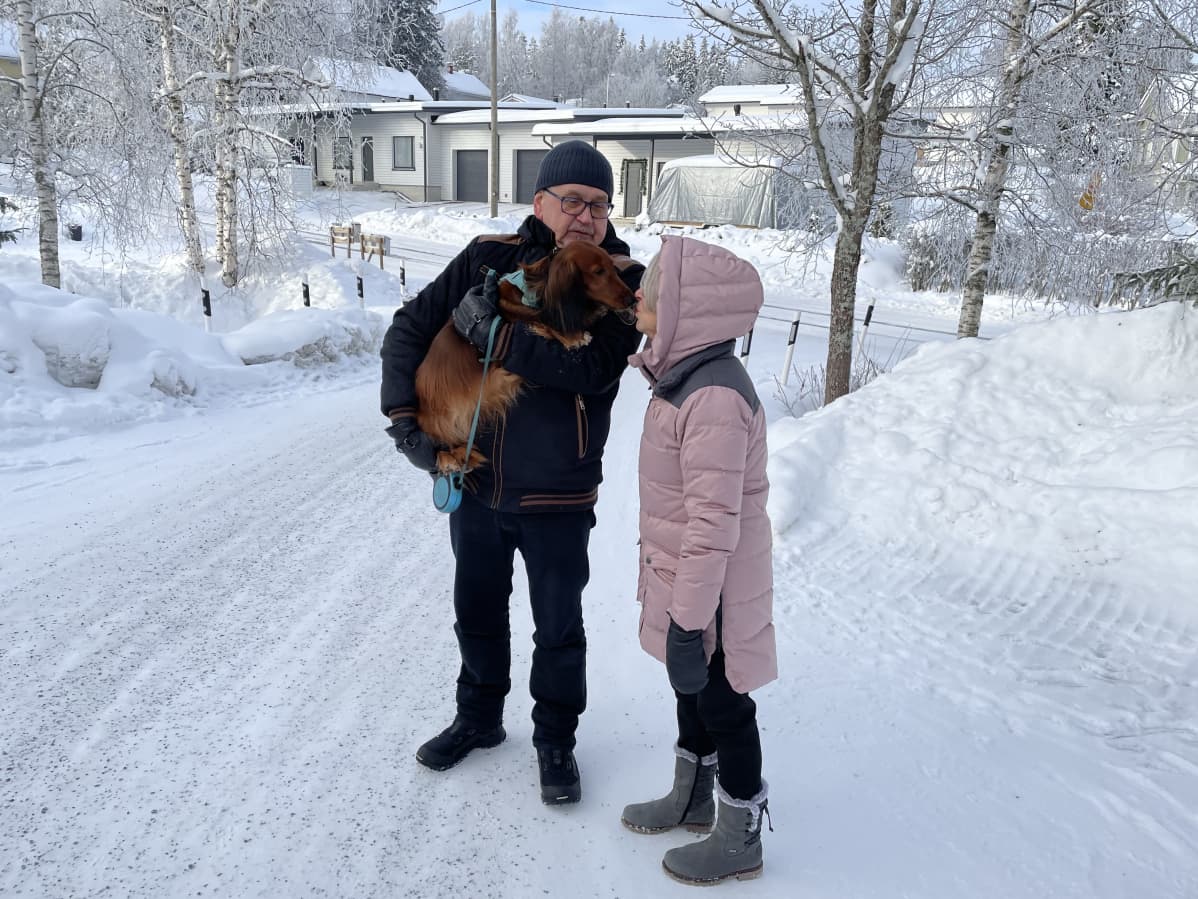 Kaksi ihmistä talvisessa maisemassa pihalla. Miehellä on sylissään koira, nainen antaa koiralle pusun.