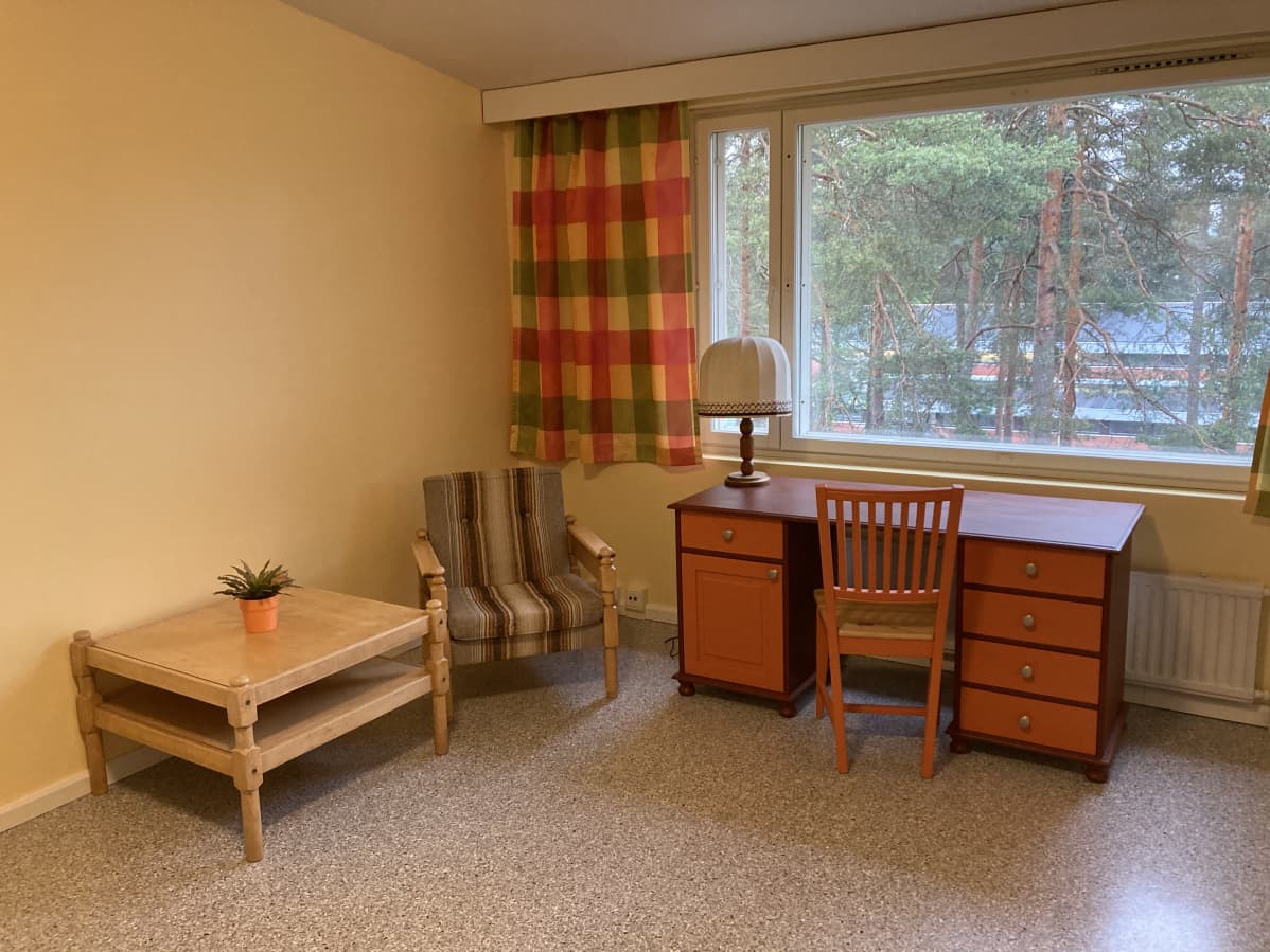 Ksaon hostellihuone. Pöytä, nojatuoli, työpöytä.