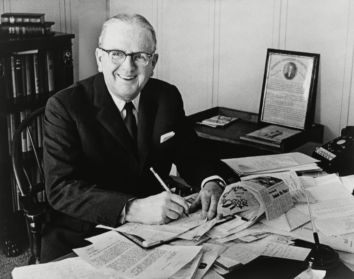 Norman Vincent Peale istuu hymyilevänä sotkuisen työpöydän ääressä mustavalkoisessa kuvassa.