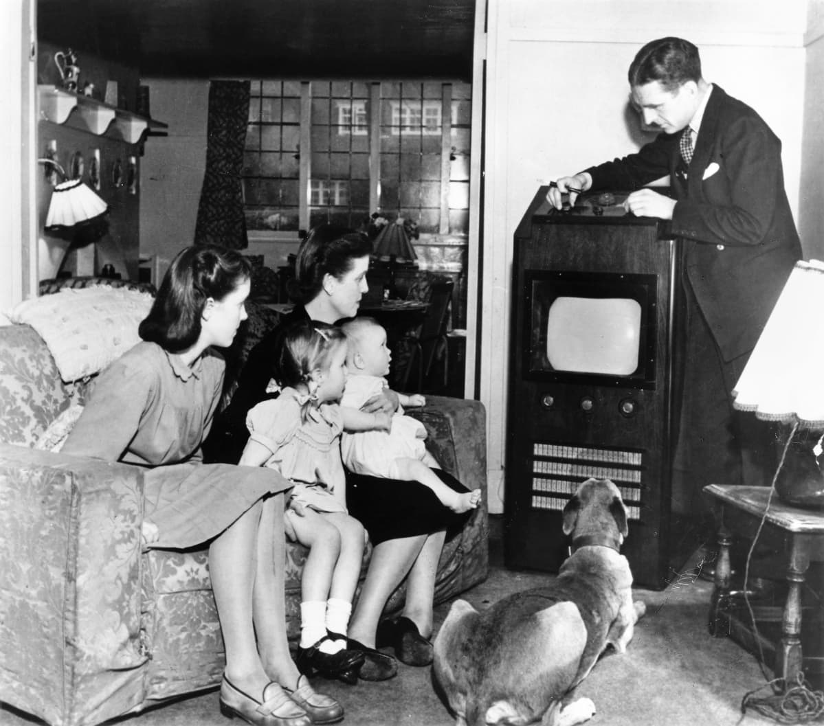 Mustavalkokuvassa perhe katsoo televisiota. Äiti vauva sylissään ja kaksi tytärtä istuu sohvalla, koira makaa lattialla ja perheenisä säätää televisiovastaanotinta.