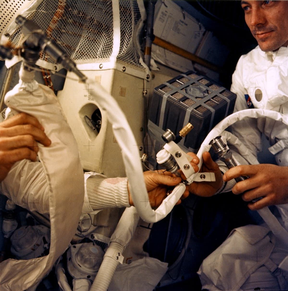 Laitteita ja kaksi astronauttia ahtaassa kuumoduulissa. Vasemmalla olevan astronautin kasvot eivät näy.