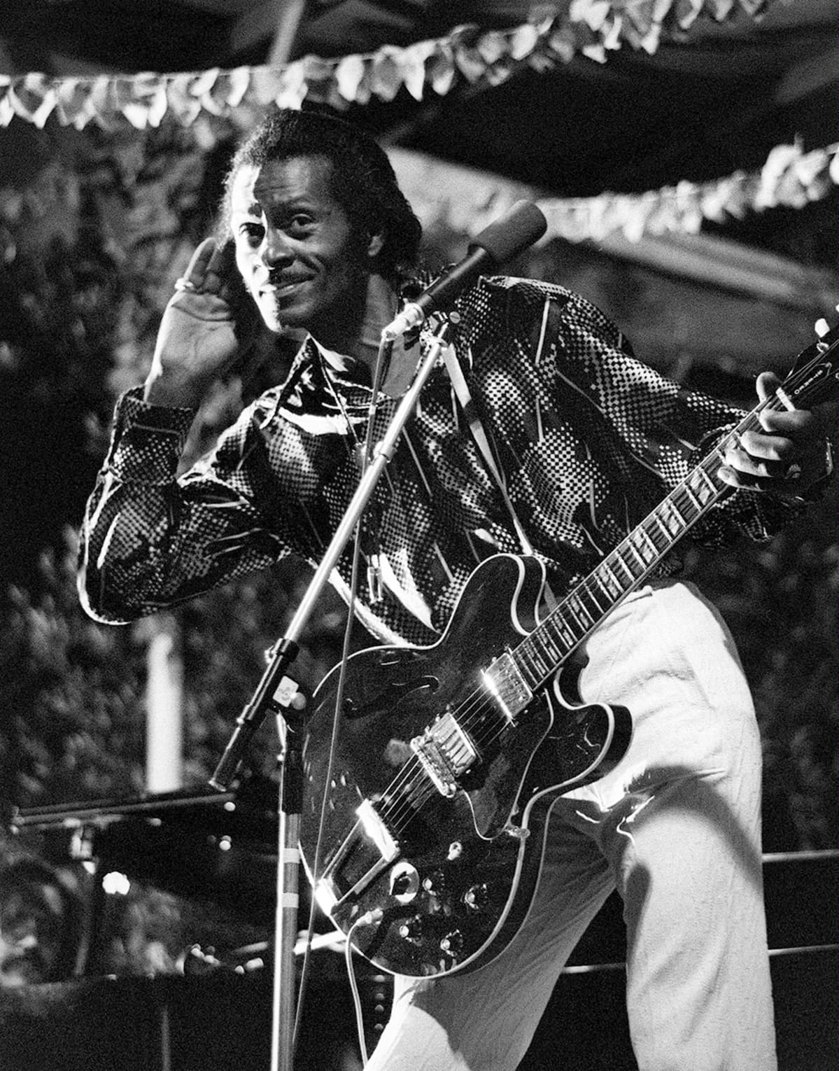 Chuck Berry kuuntelee yleisön äännehdintää oikea käsi oikealla korvallaan ja katsoo lempeän leikkimielisesti valokuvaajaa kohti.