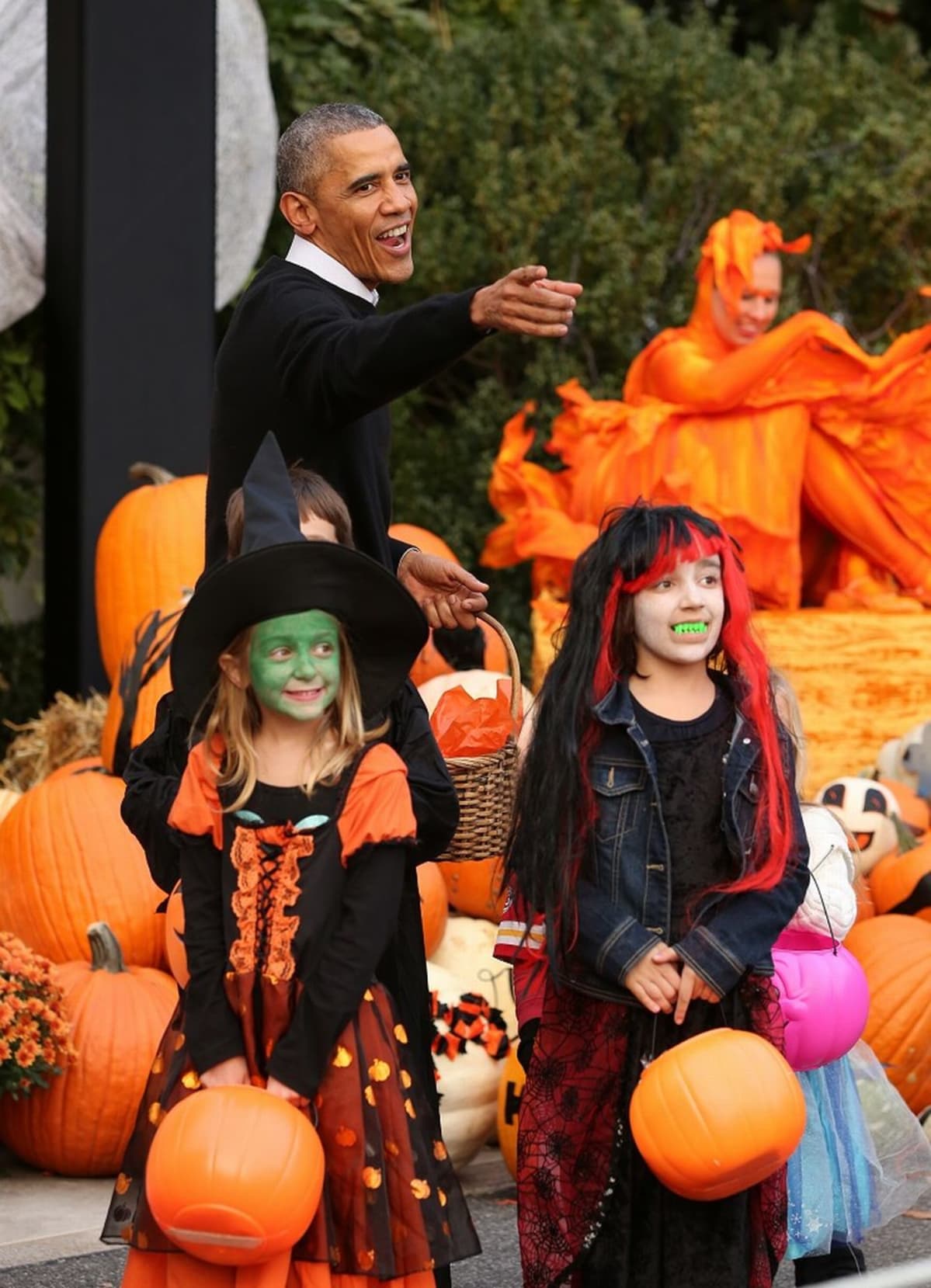 Kaksi noidiksi pukeutunutta pikkutyttöä ja presidentti Barack Obama, jolla on kädessään pajukori. Ympärillä kurpitsoja.
