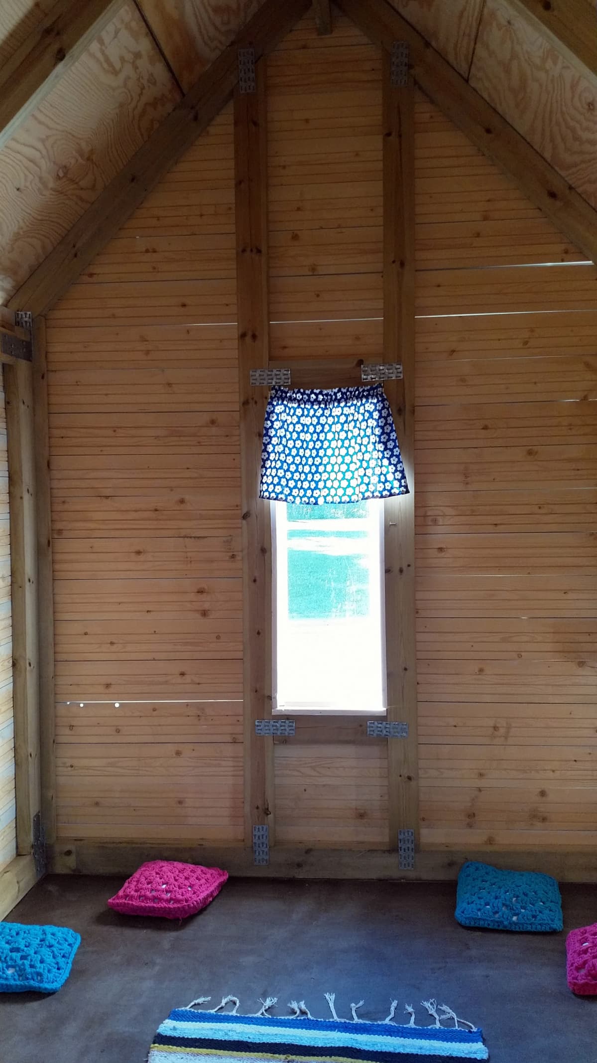 Kuvassa leikkimökki sisältä. Lattialla on neljä virkattua tyynyä ja räsymatto. Ikkunassa on verho.