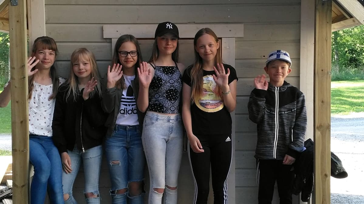 Kuvassa kuusi oppilasta seisoo leikkimökin edessä ja vilkuttaa.