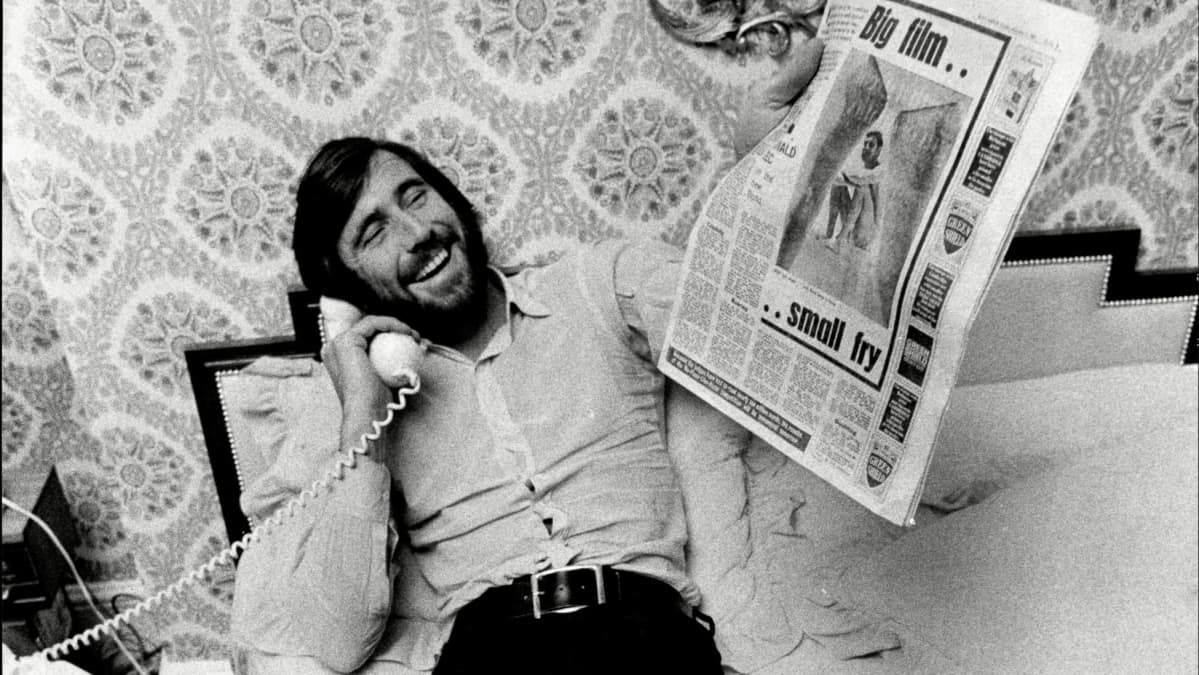 George Lazenby vanhassa valokuvassa, puhelin kädessä ja sanomalehti toisessa. Kuva dokumentista Becoming Bond.