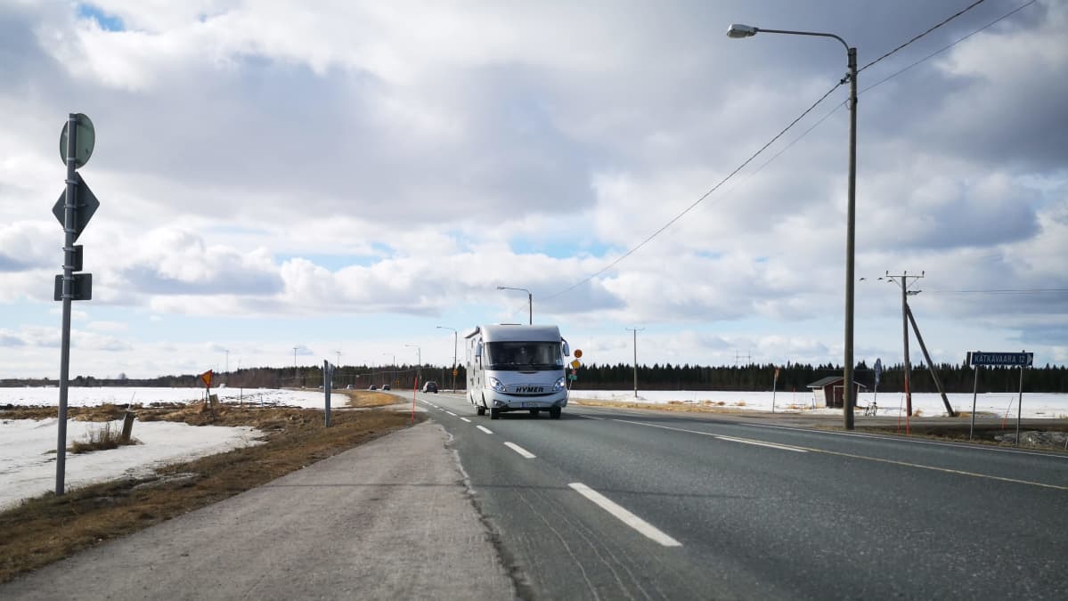 Matkailuauto lähestyy sulaa asvalttitietä pitkin, ympärillä näkyy lumisia peltoja.