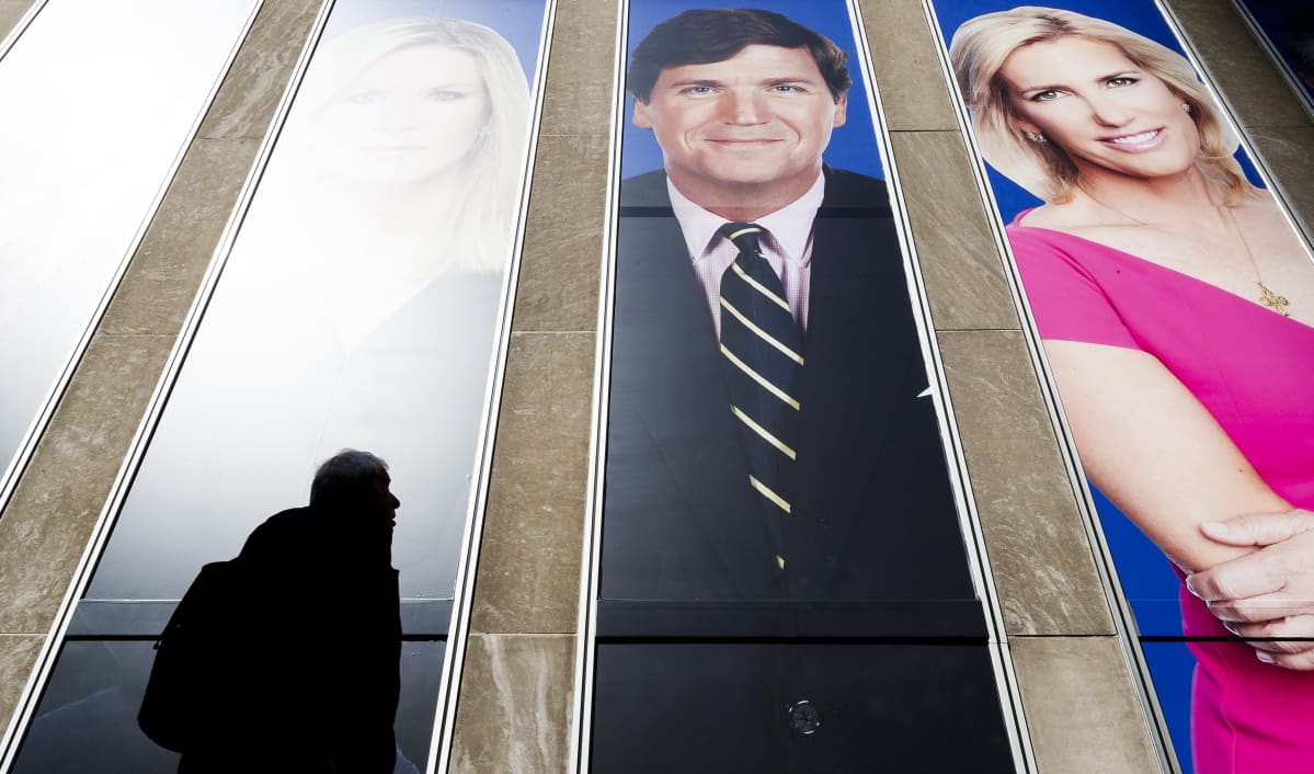 Fox Newsin ankkureita mainoksessa, mm. Tucker Carlson, mies kävelee mainoksen ohi.