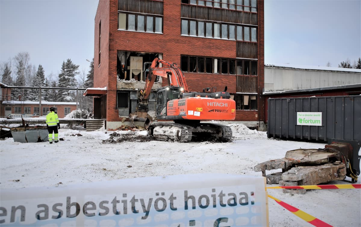 Kaivinkone hajottaa tiiliseinää, kaksi henkilöä tarkkailee purkua