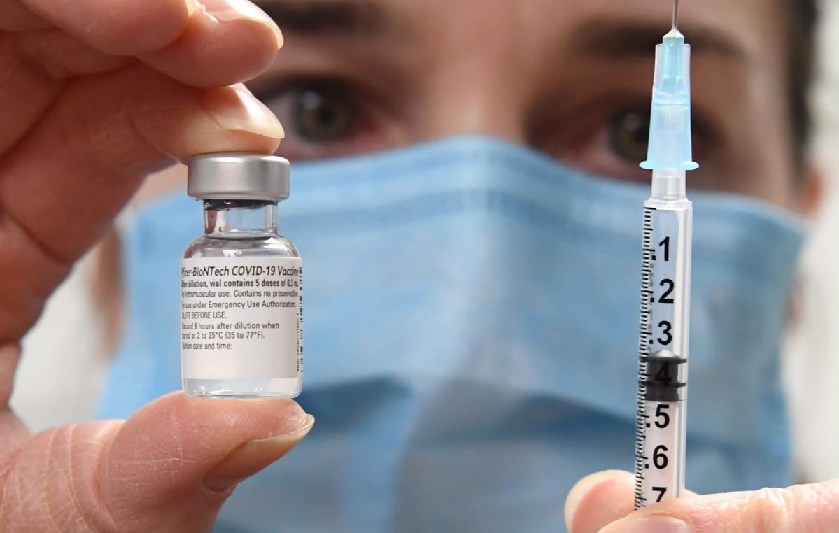 I bakgrunden syns ett ansikte med munskydd. Kameran fokuserar på en liten glasflaska med coronavaccin och en spruta personen håller i.