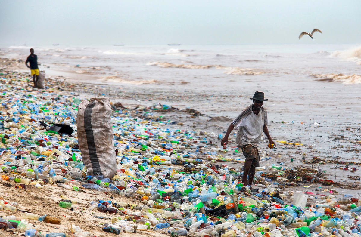 Kaksi miestä kerää purkkeja säkkeihin rannalla, joka on täynnä kirjavaa muovijätettä. 