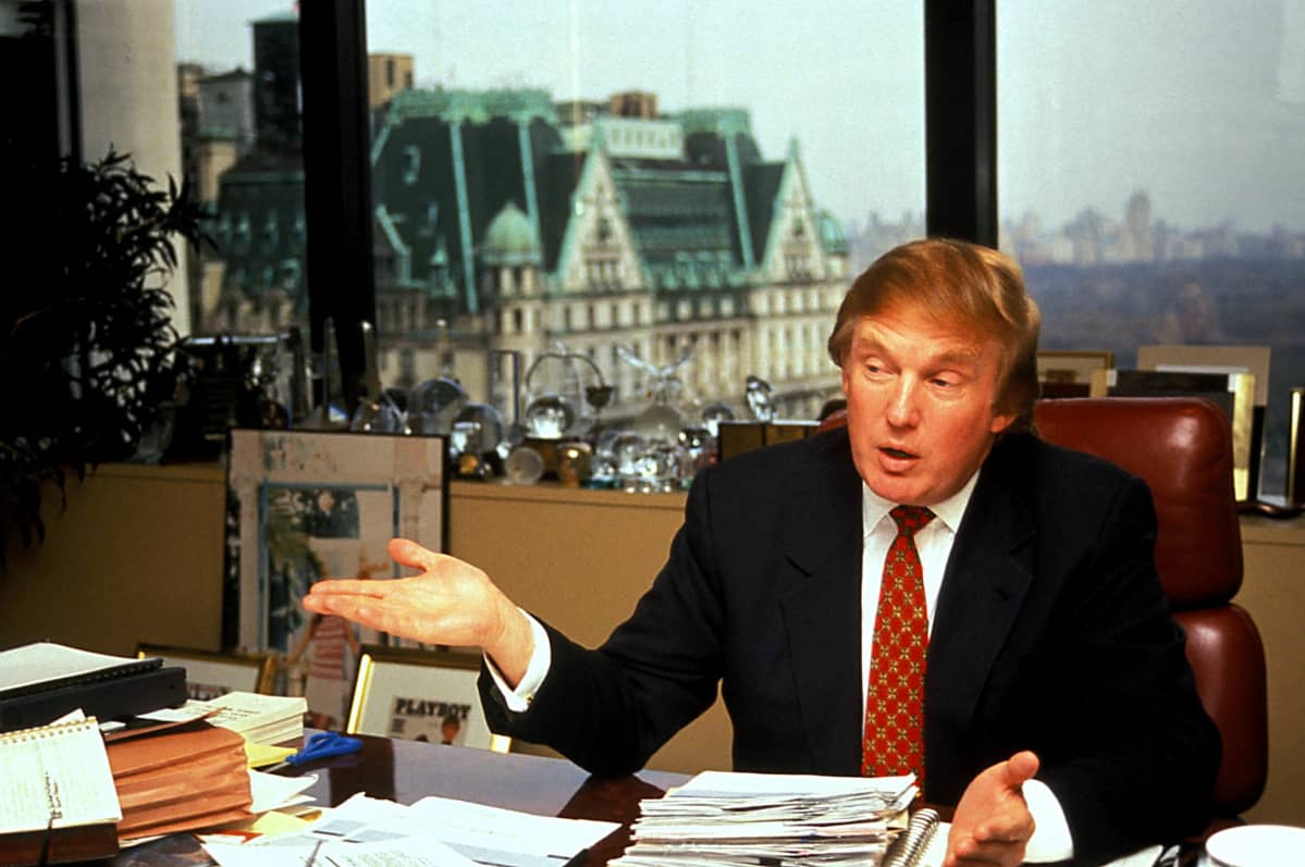 Donald Trump istuu työpöytänsä ääressä ehkä 1990-luvulla. Takana ikkunasta aukeaa kaupunkimaisema.