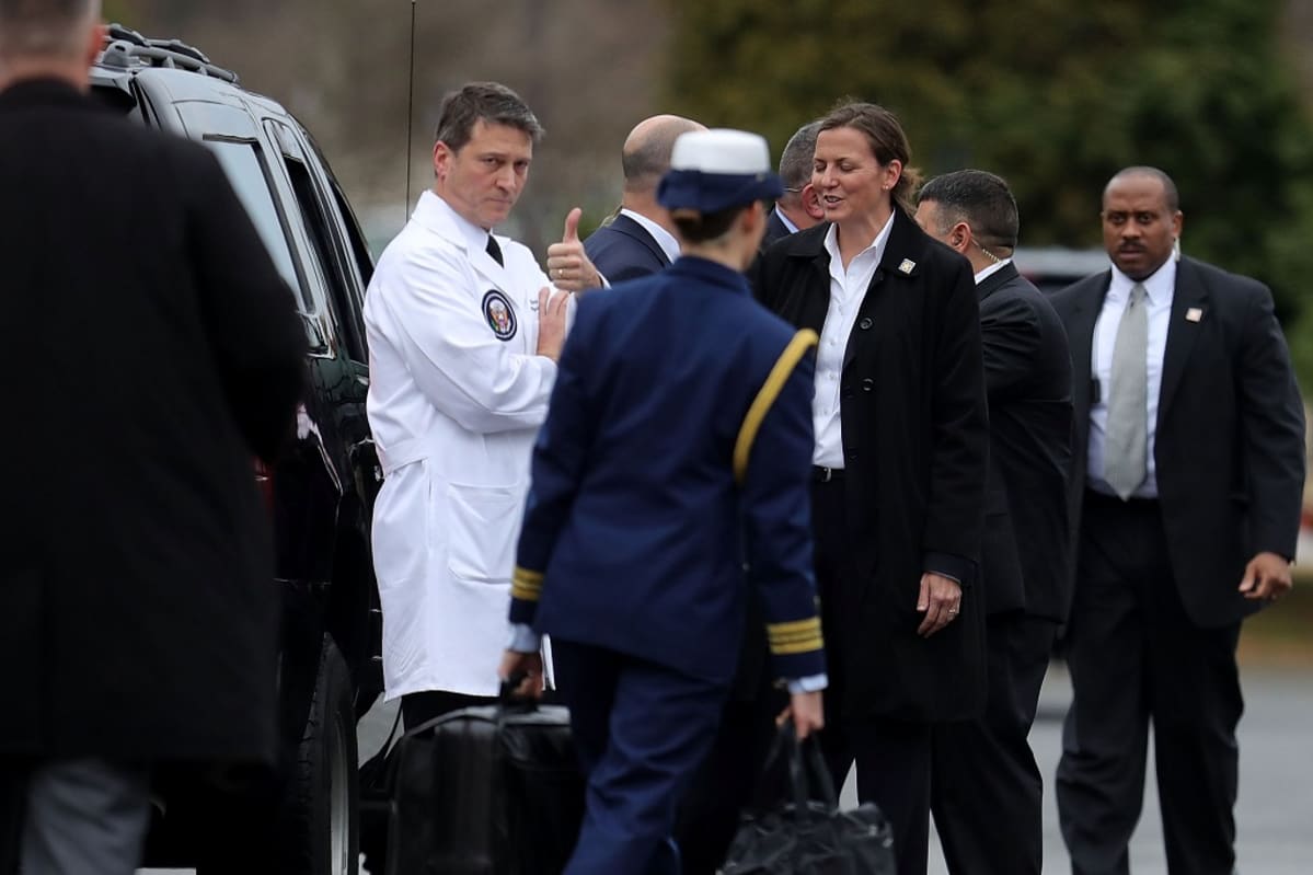 Ronny Jackson seisoo valkoisessa lääkärintakissa mustaa autoa vasten kädet ristissä, katsoo kameraan ja nostaa peukaloa. Ympärillä parveilee muuta Valkoisen talon henkilökuntaa.