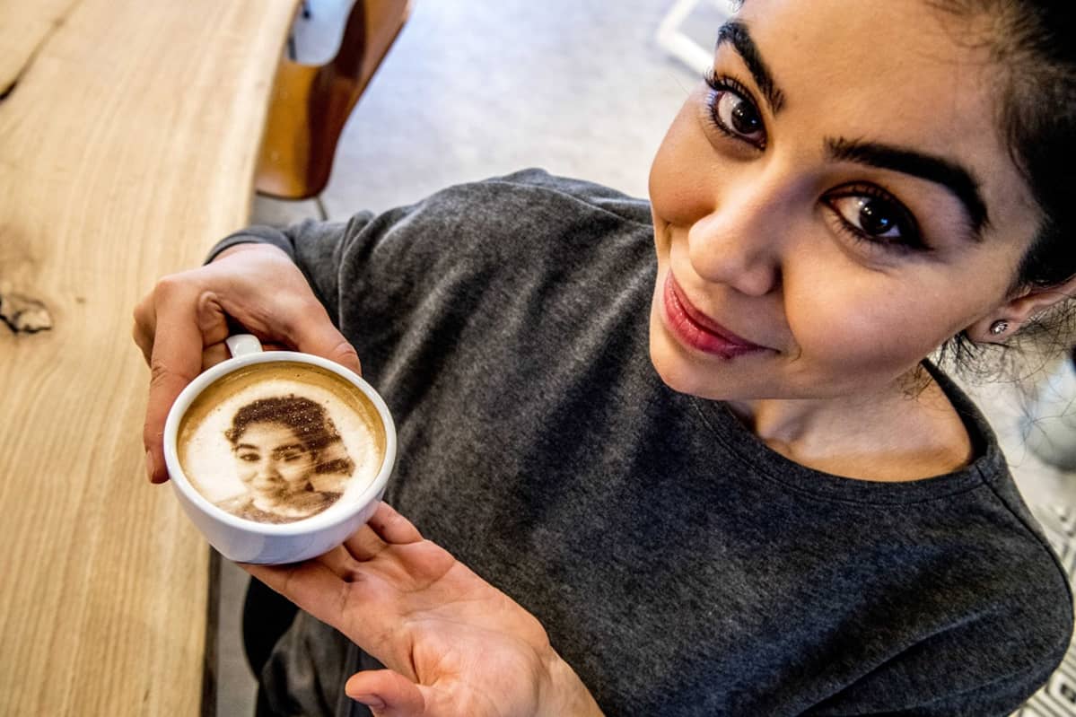 Nainen pitelee kuppia, jonka kahvin kintaan on muotoiltu vaahdosta hänen selfie-kuvansa.