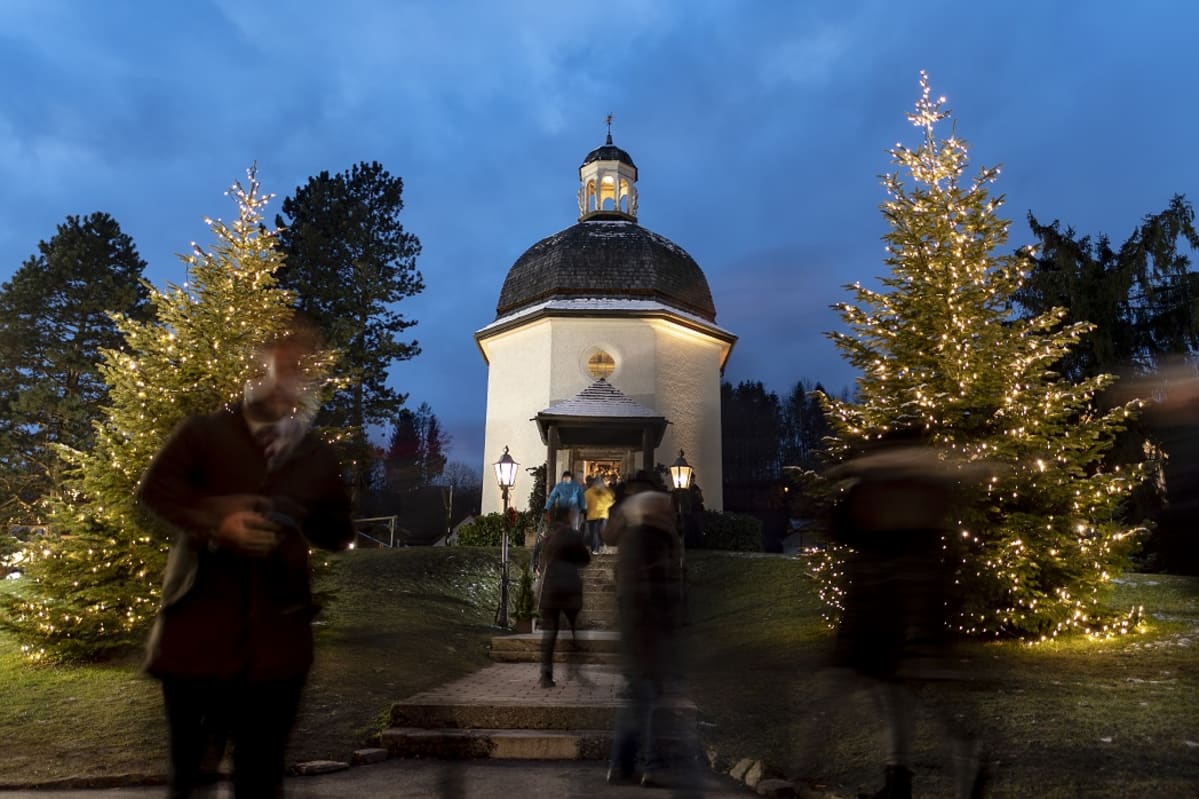 Kaksi valaistua joulukuusta sininen taivasta ja valkoinen kappeli.