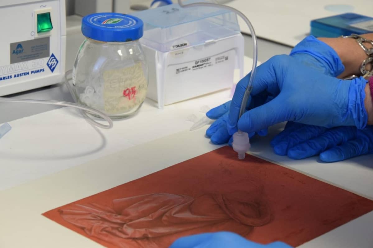 Naisen hansikoidut kädet liikkuttavat anturia piirroksen pinnalla. Taustalla laboratoriopöydän laitteistoa.