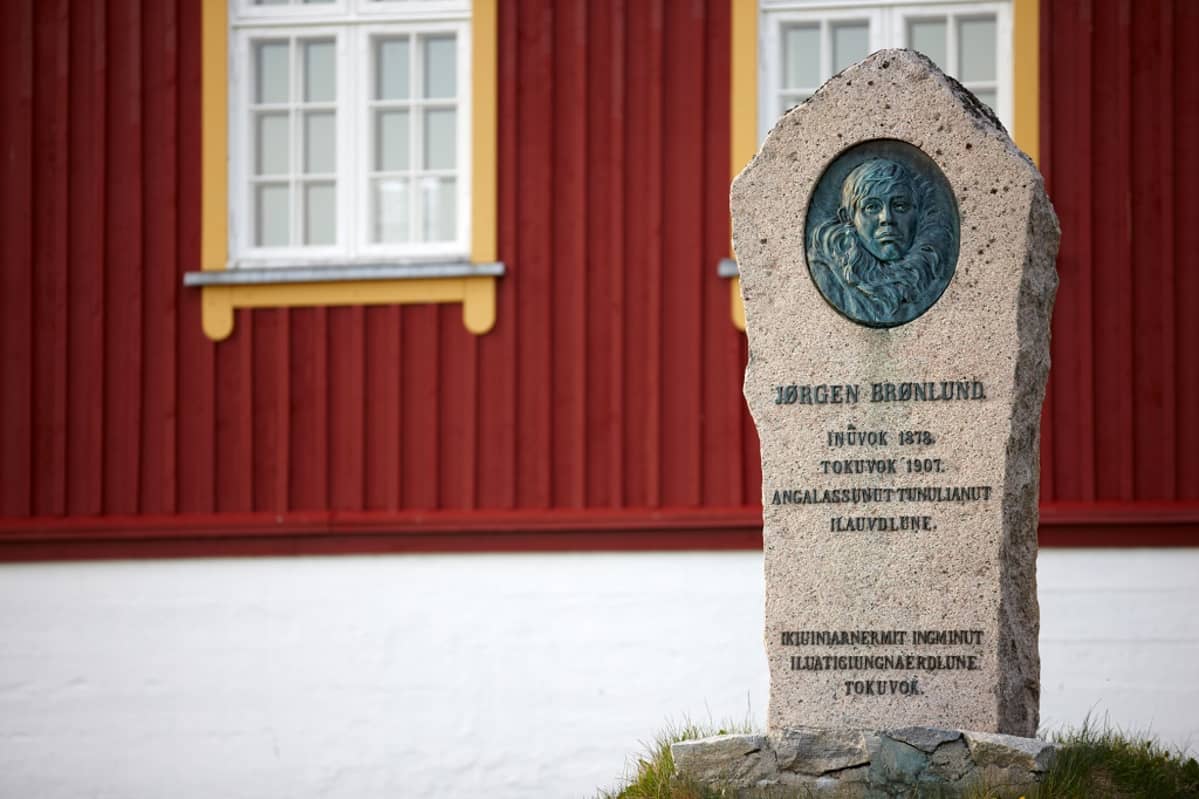 Muistokivi punaisen puurakennuksen edessä. Kivessä on Jørgen Brønlundin kasvokuva ja synnyin- ja kuolinpaikat ja -vuodet: Inuvok 1878, Tokuvok 1907.